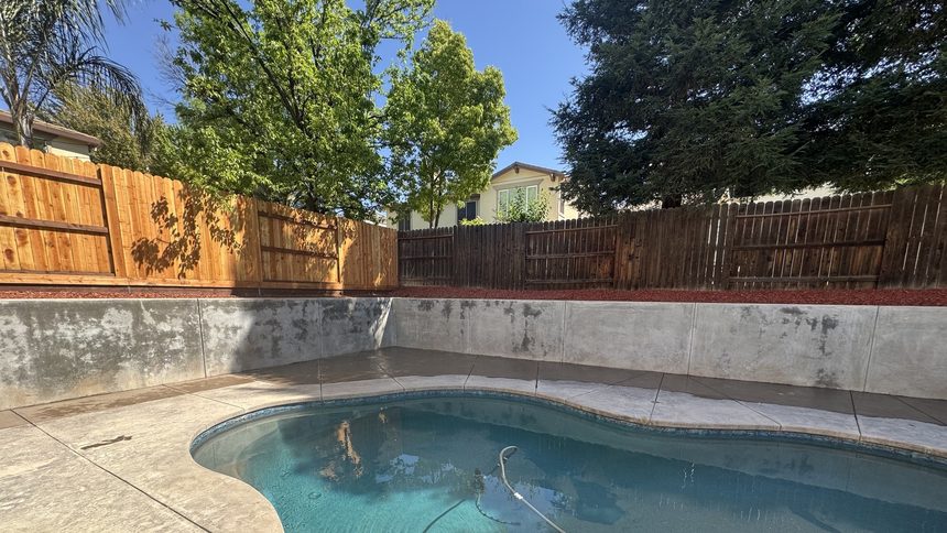 A poured concrete retaining wall running the full width of a yard, holding a bark bed back off a stamped pool deck.