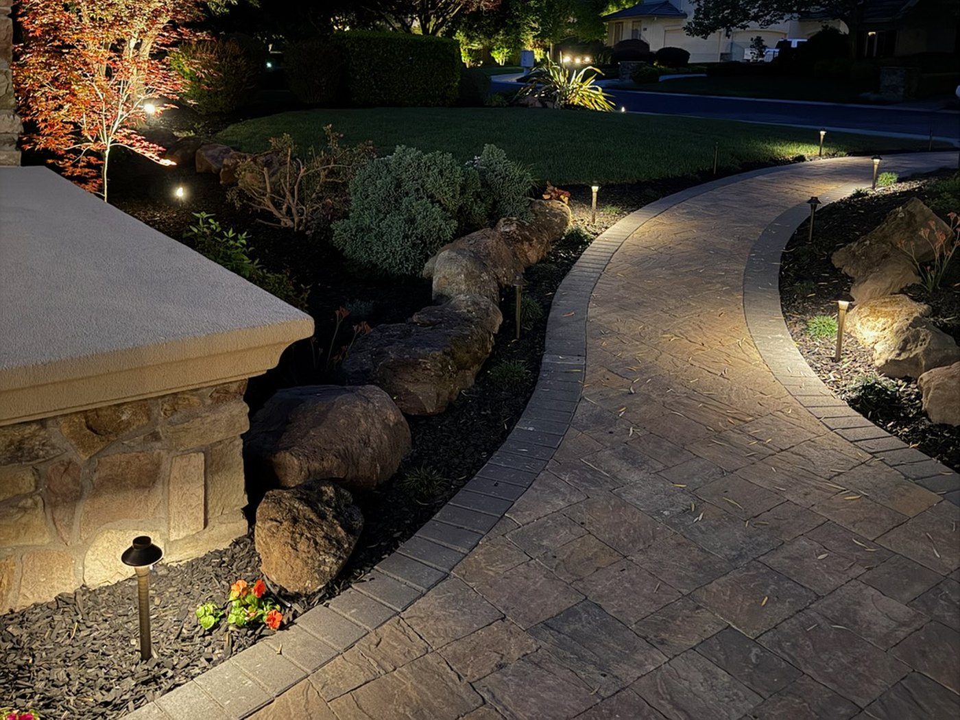A paver walk lit along its length at dusk, running between boulder-edged planting beds.